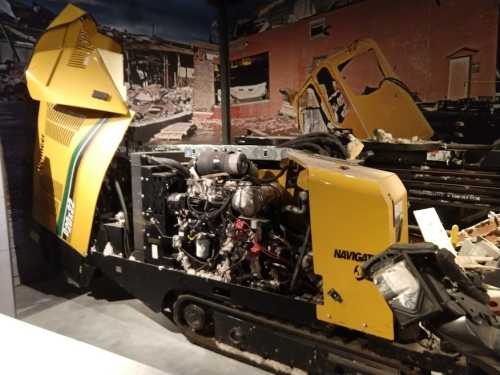 A damaged yellow snow groomer displayed in a museum, showcasing its exposed engine and surrounding debris.