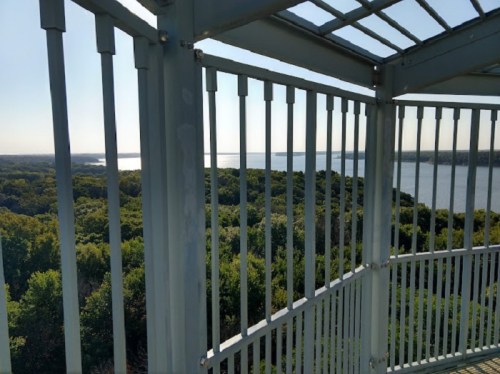 A view from a lookout tower, showing lush green trees and a shimmering lake in the distance under a clear sky.