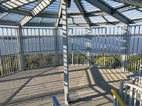 View from inside a metal observation tower, featuring a circular floor and railings, overlooking a river and trees.