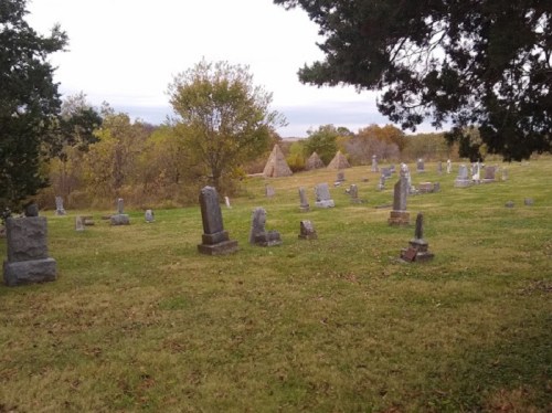 A grassy cemetery with numerous gravestones and trees, set against a cloudy sky.