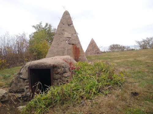 Two pyramid-shaped stone structures on a grassy hill, surrounded by sparse trees and cloudy skies.