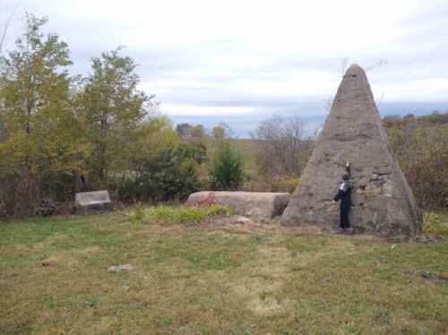 A person stands beside a large stone structure in a grassy area, surrounded by trees and a cloudy sky.