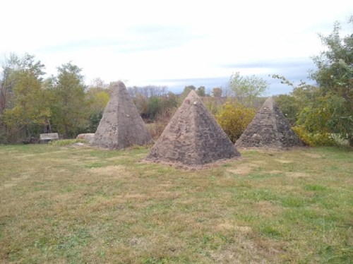 Three stone pyramids in a grassy area, surrounded by trees and a cloudy sky.