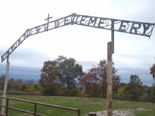 Rusty metal archway with the word "Cemetery" and a cross, set against a cloudy sky and autumn trees.