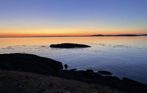A serene sunset over calm waters, with silhouettes of rocks and distant hills against a colorful sky.