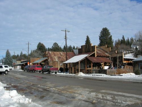 A rustic town street with wooden buildings, snow on the ground, and parked trucks under a cloudy sky.