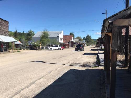 A quiet, empty street in a small town with wooden buildings and parked cars under a clear blue sky.