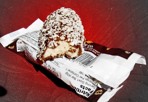 A partially eaten ice cream bar coated in chocolate and coconut flakes, resting on its wrapper.