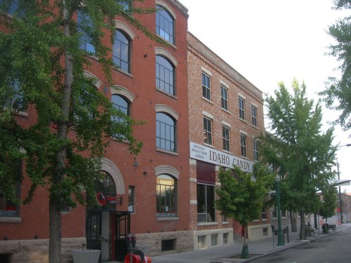 Historic brick buildings with large windows, featuring a sign for "Idaho Candy," lined by trees on a quiet street.