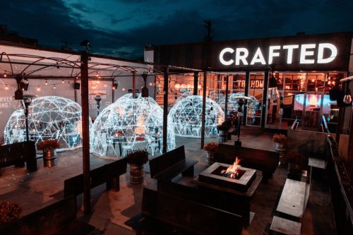 Outdoor dining area with illuminated igloos, a fire pit, and a sign reading "CRAFTED" against a twilight sky.