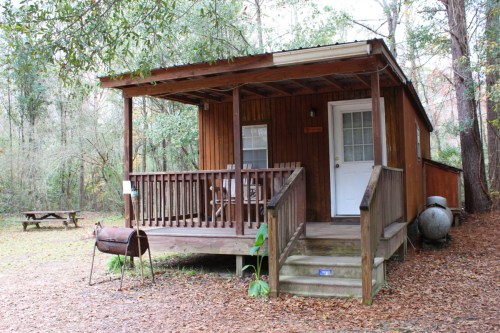 A small wooden cabin with a porch, surrounded by trees and a picnic table nearby.
