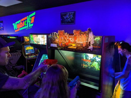 Two players aim at a "House of the Dead 2" arcade game in a colorful gaming arcade. Neon lights illuminate the scene.