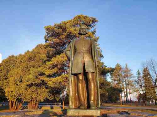 A bronze statue of a soldier stands in a park, surrounded by trees under a clear blue sky.