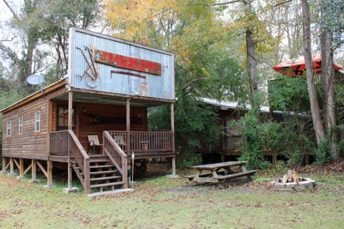 A rustic wooden cabin with a sign, surrounded by trees, featuring a picnic table and a fire pit in the yard.