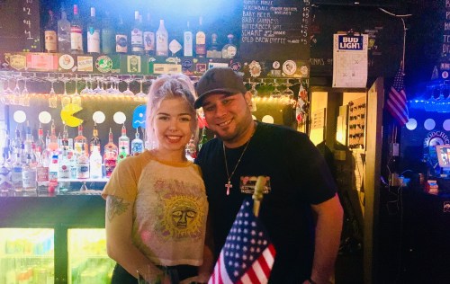 A smiling woman and man pose together in a bar, with bottles and an American flag in the background.