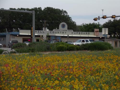 A roadside view of Hill Country Cupboard with colorful wildflowers in the foreground and traffic lights in the background.