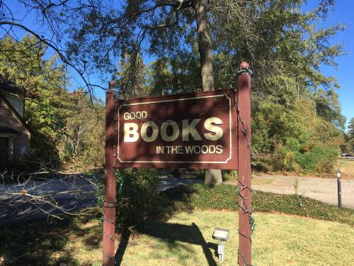 Sign reading "Good Books in the Woods" surrounded by trees and greenery on a sunny day.