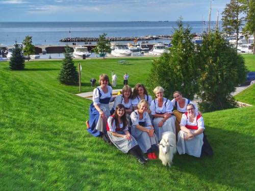A group of women in traditional dresses pose on a grassy hill by the water, with boats and trees in the background.