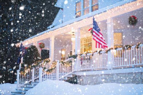 A cozy house decorated for the holidays, with snow falling and American flags displayed on a snowy porch.