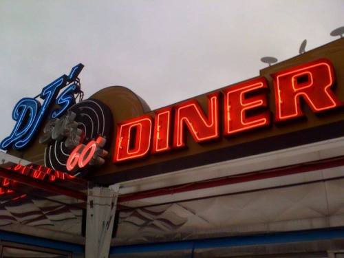 Neon sign of DJ's 60's Diner, featuring a retro vinyl record design and bright red lettering against a cloudy sky.