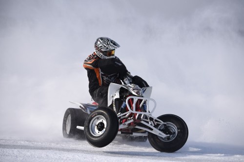 A rider on a quad bike speeds through a snowy landscape, kicking up a cloud of snow behind them.