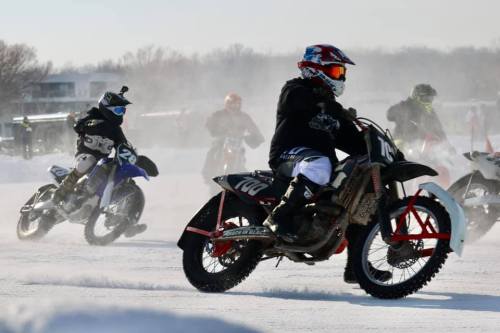 Motorcyclists racing on a snowy track, kicking up snow and dust in a winter motorsport event.