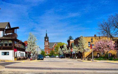 A charming town square with blooming trees, historic buildings, and a church steeple under a clear blue sky.