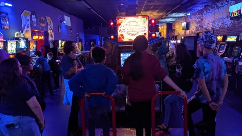A group of people gathered around an arcade game in a vibrant gaming venue filled with pinball machines.