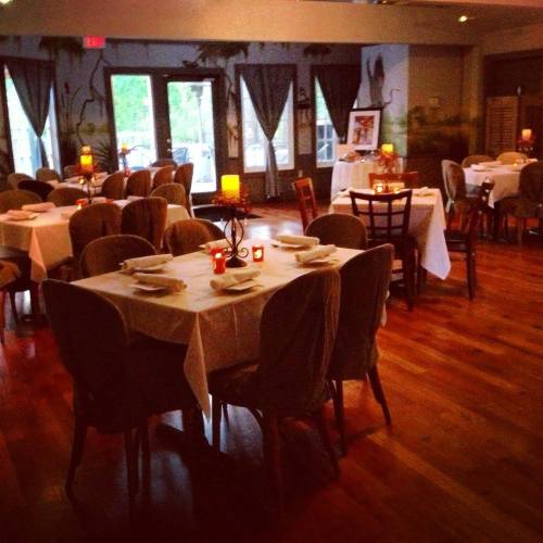 A cozy restaurant interior with tables set for dining, warm lighting, and large windows overlooking greenery.