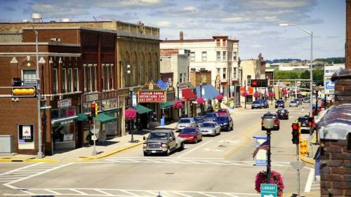 A bustling small-town street with shops, cars, and colorful flower baskets under a partly cloudy sky.