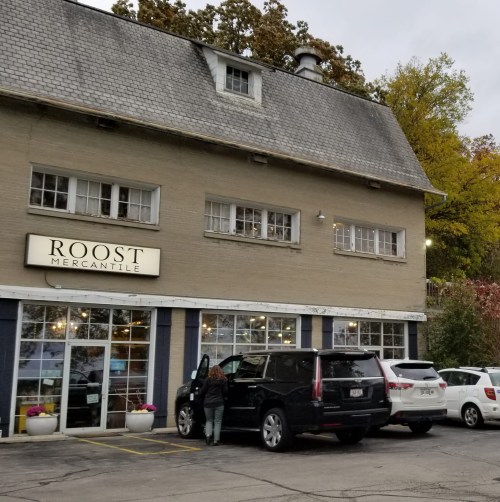 A storefront labeled "Roost Mercantile" with large windows, a gray exterior, and a black SUV parked outside.