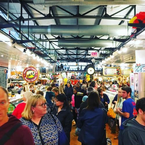 A bustling market interior filled with people browsing various food stalls and shops under bright lights.