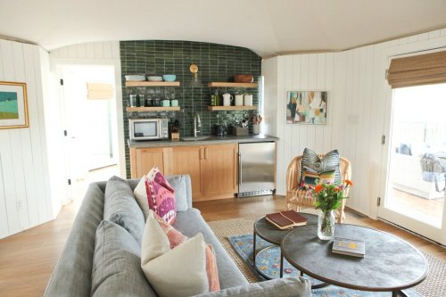 Cozy living space with a gray sofa, wooden shelves, and a small kitchen area featuring green tiles and natural light.