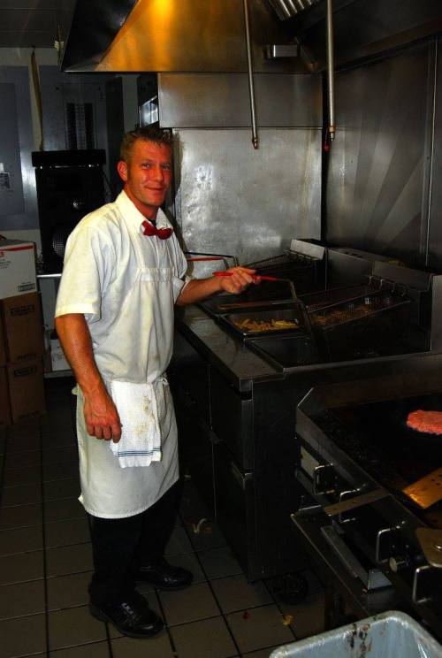 A chef in a white uniform and red bow tie stands at a grill in a kitchen, cooking with a spatula.