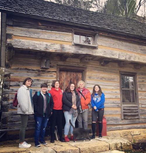 A group of six women stands in front of a rustic wooden cabin, smiling together outdoors.