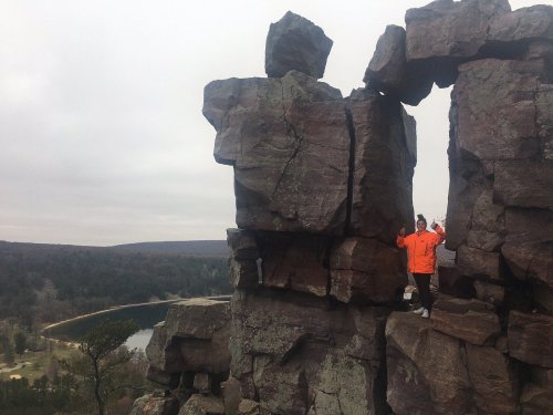 A person in an orange jacket stands between two large rock formations overlooking a scenic landscape.