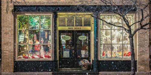A festive toy store window decorated for winter, with colorful displays and snow falling outside.