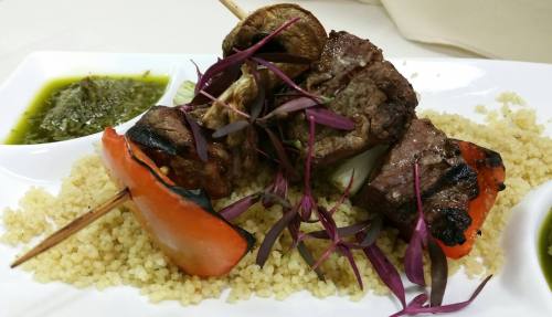Grilled meat skewers with vegetables on a bed of couscous, garnished with herbs and sauces on the side.
