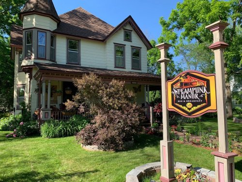 A charming Victorian-style house with a colorful sign reading "Steampunk Manor Bed & Breakfast" in a lush garden.