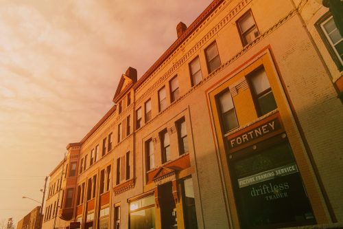 Historic brick buildings with a warm sunset glow, featuring storefronts and a cloudy sky above.