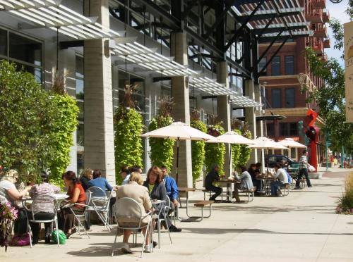 Outdoor café scene with people sitting at tables under umbrellas, surrounded by greenery and modern architecture.