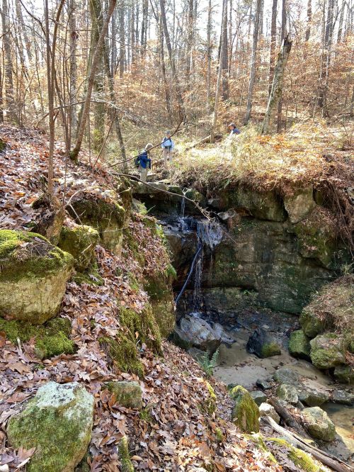 Two hikers explore a rocky area with a small waterfall, surrounded by autumn foliage and trees in a forested setting.