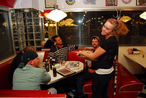 A waitress smiles while serving drinks to a group of four men at a retro diner booth.