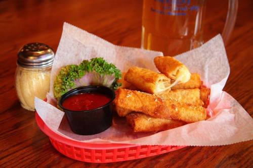 A basket of crispy spring rolls with marinara sauce, lettuce, and a shaker of seasoning, alongside a mug of beer.