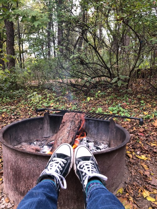 A pair of feet in sneakers resting near a campfire with a log burning, surrounded by autumn foliage.