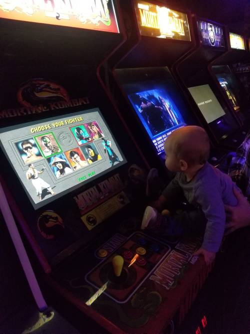 A baby sits at an arcade machine, focused on the "Mortal Kombat" game screen displaying character selection.