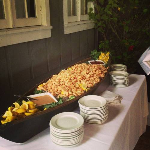 A large platter of shrimp with lemon slices, accompanied by dipping sauces and stacked white plates on a table.