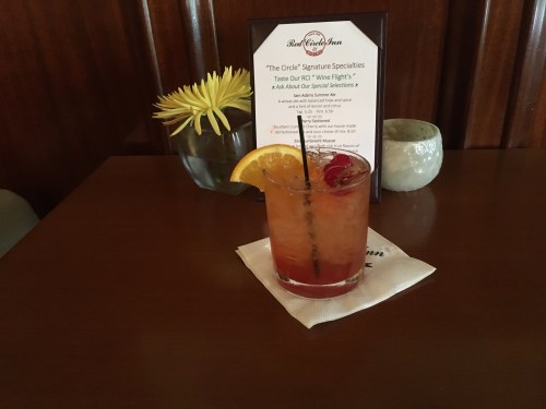 A colorful cocktail with an orange slice and cherry, placed on a table next to a menu and a flower vase.