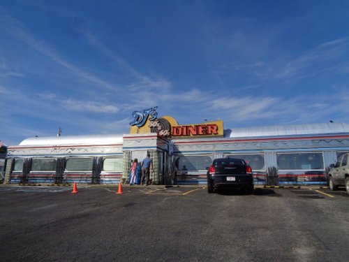 A retro diner with a silver exterior and colorful signage, featuring a parking lot and two people outside.