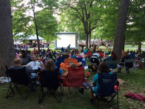 A crowd sits on lawn chairs in a park, watching a movie on a large outdoor screen surrounded by trees.